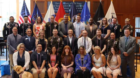 A large group of people pose in front of U.S. military flags.