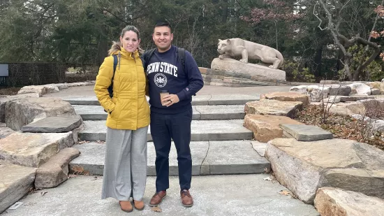 A man and woman stand in front of a rocky area and lion sculpture.