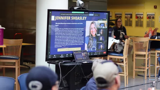 A crowd of people view a woman presenting on a TV screen.