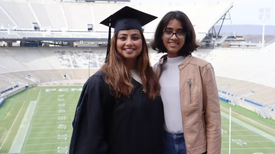 A woman wearing a black graduation cap and gown stands with another woman in front of an empty stadium.