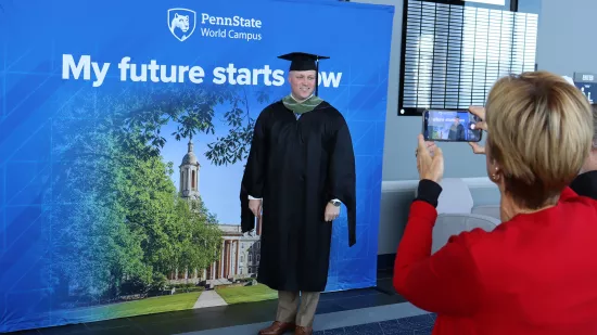 A woman takes a photo of a man in front of a Penn State backdrop.