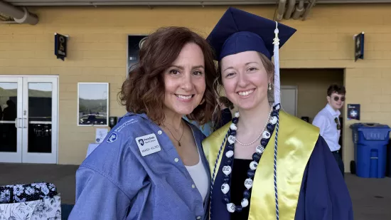 Two women, one wearing a blue graduation cap and gown, pose for a photo in an outdoor area.