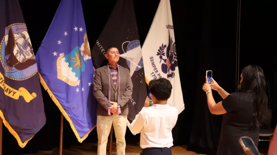 Two people take photos of a man standing in front of a group of flags.