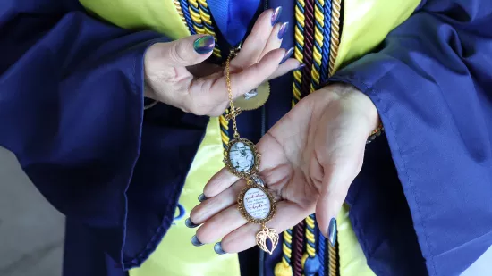 A woman wearing a graduation gown holds a small locket with a photo of a loved one.