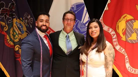 Two men wearing suits and a woman wearing a white dress pose for a photo in front of a group of flags.