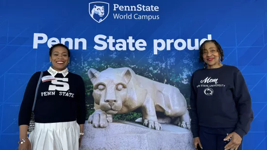 Two women wearing Penn State apparel pose in front of a Penn State photo background.