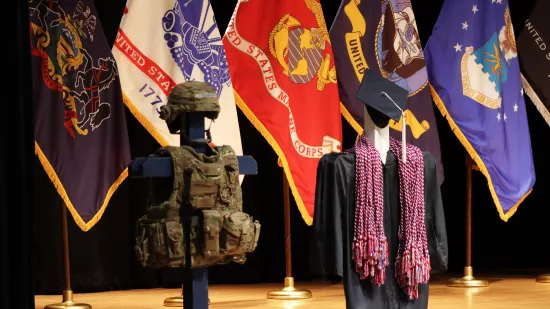 Military body armor and a blue graduation cap and gown are displayed in front of a group of flags.