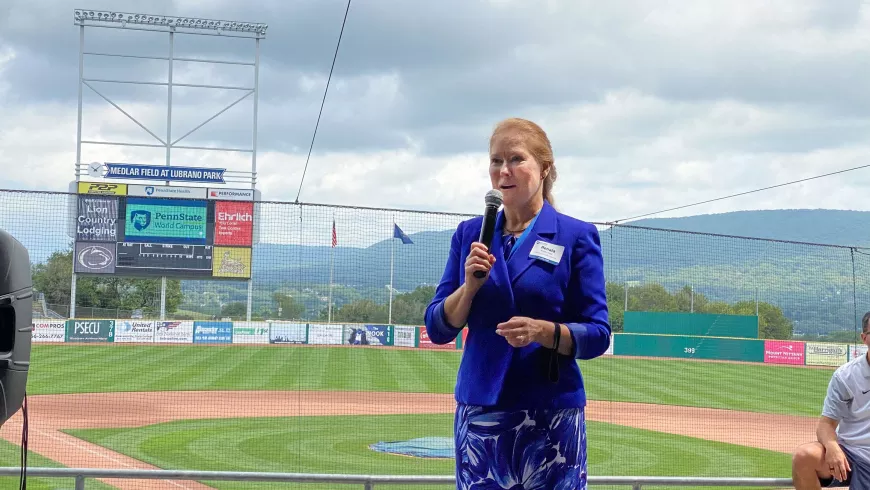 Renata Engel addresses the crowd at the graduation celebration from a platform with a baseball diamond in the background.