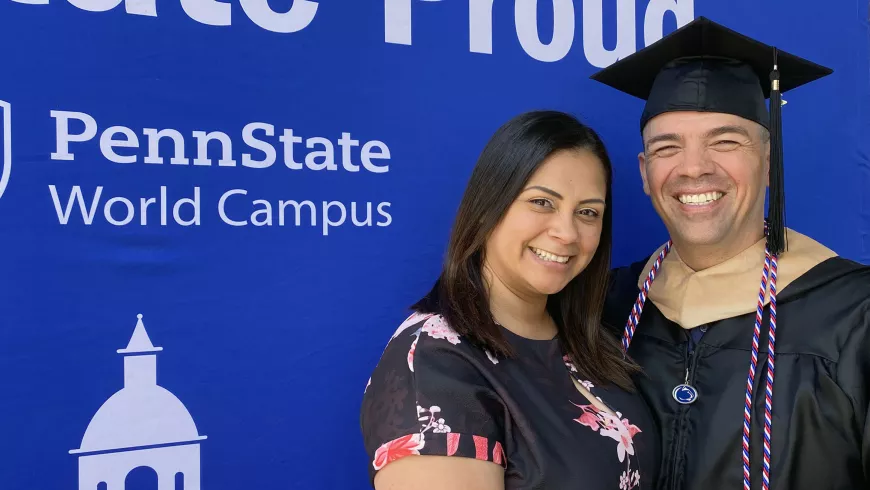 Joshua Celis is wearing a black graduation cap and gown with red, white and blue honor cords while standing with his Wife against a Penn State World Campus backdrop