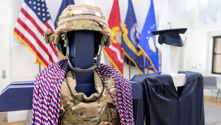 Military honor cords are draped around a military equipment on a post, which is next to a post that also has a graduation cap and gown over it.