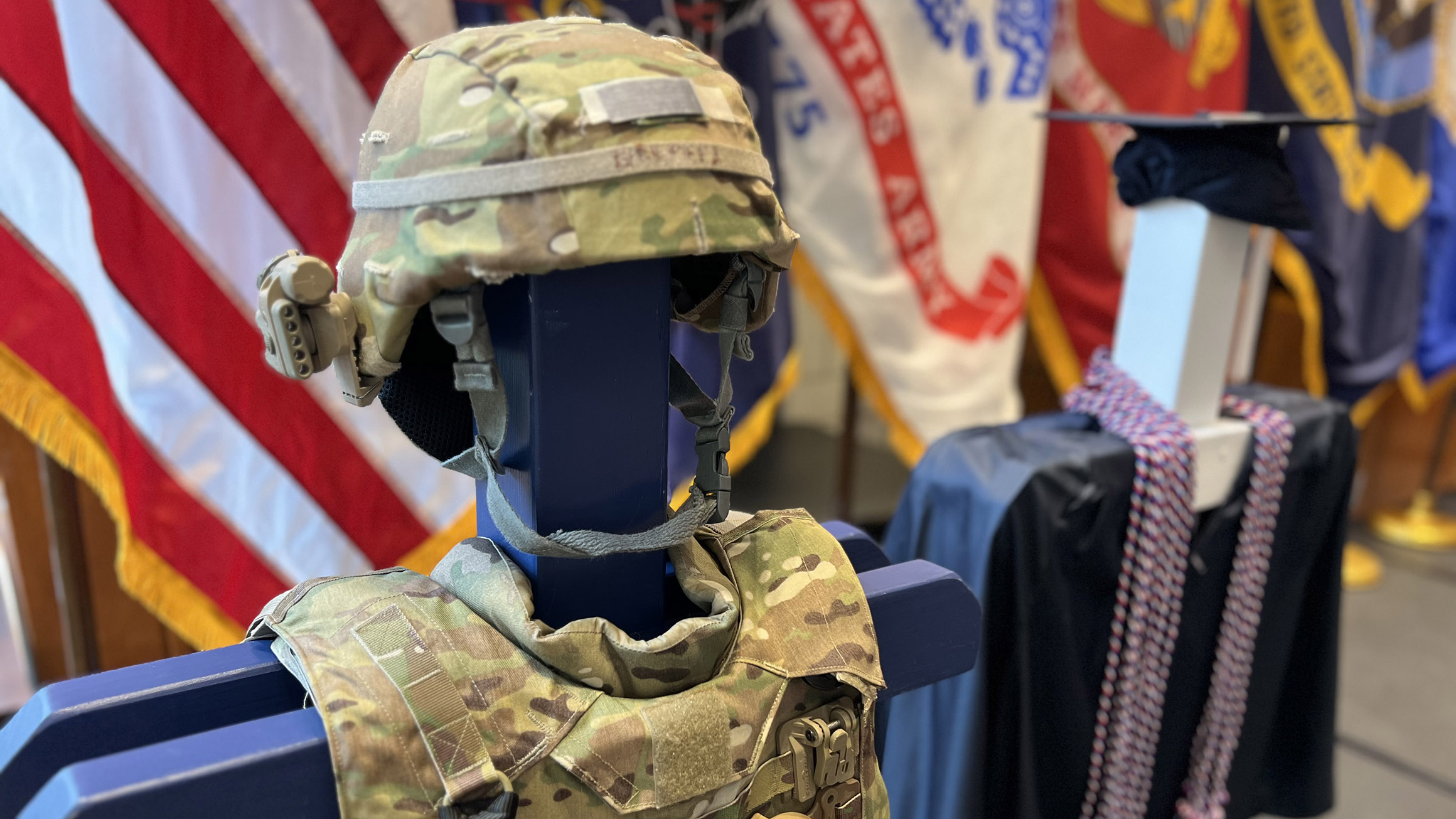 Military gear and honor cords draped around a graduation gown are shown on a dummy