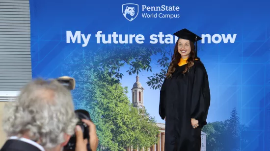 A woman wearing a black graduation cap and gown stands in front of a blue and white background. 